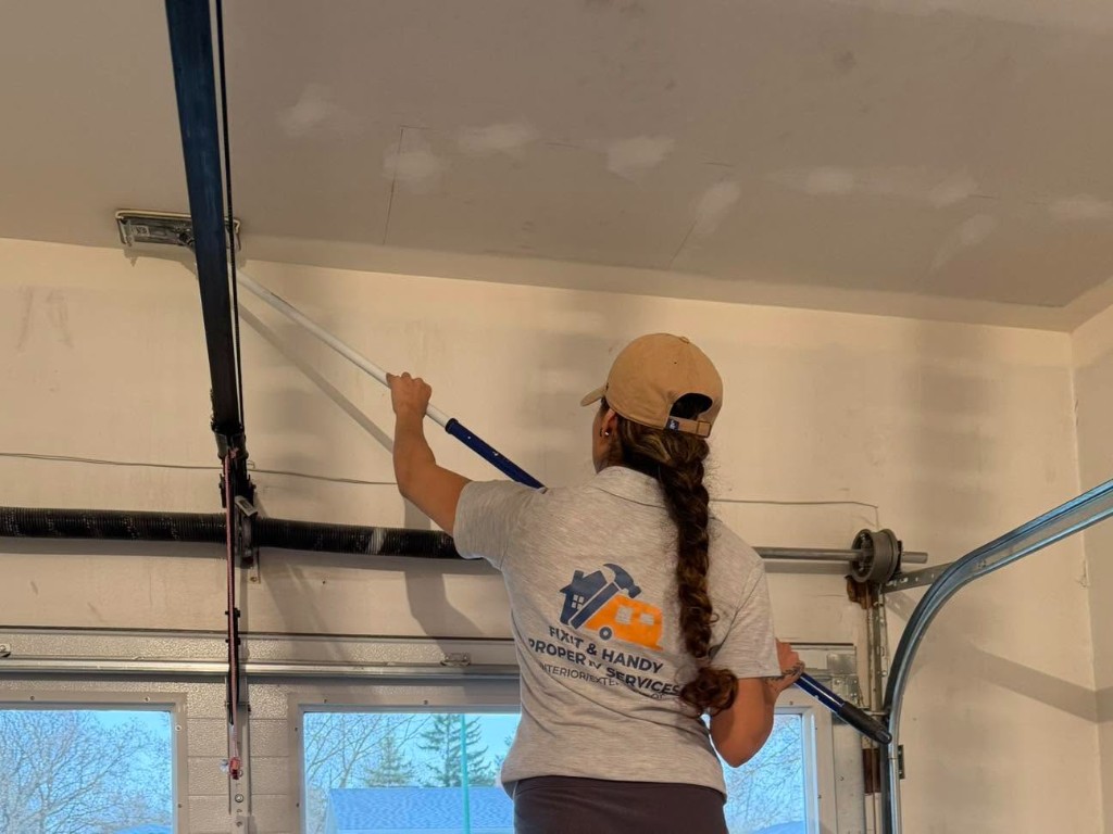 Fixit & Handy worker sanding drywall patches on a garage ceiling with pole sander.