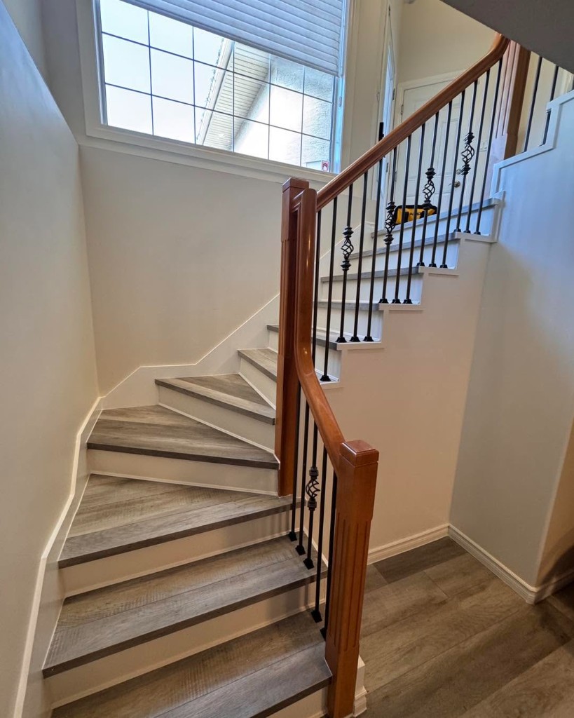 Finished interior staircase with vinyl plank flooring, white risers, and wood handrail with black metal balusters.
